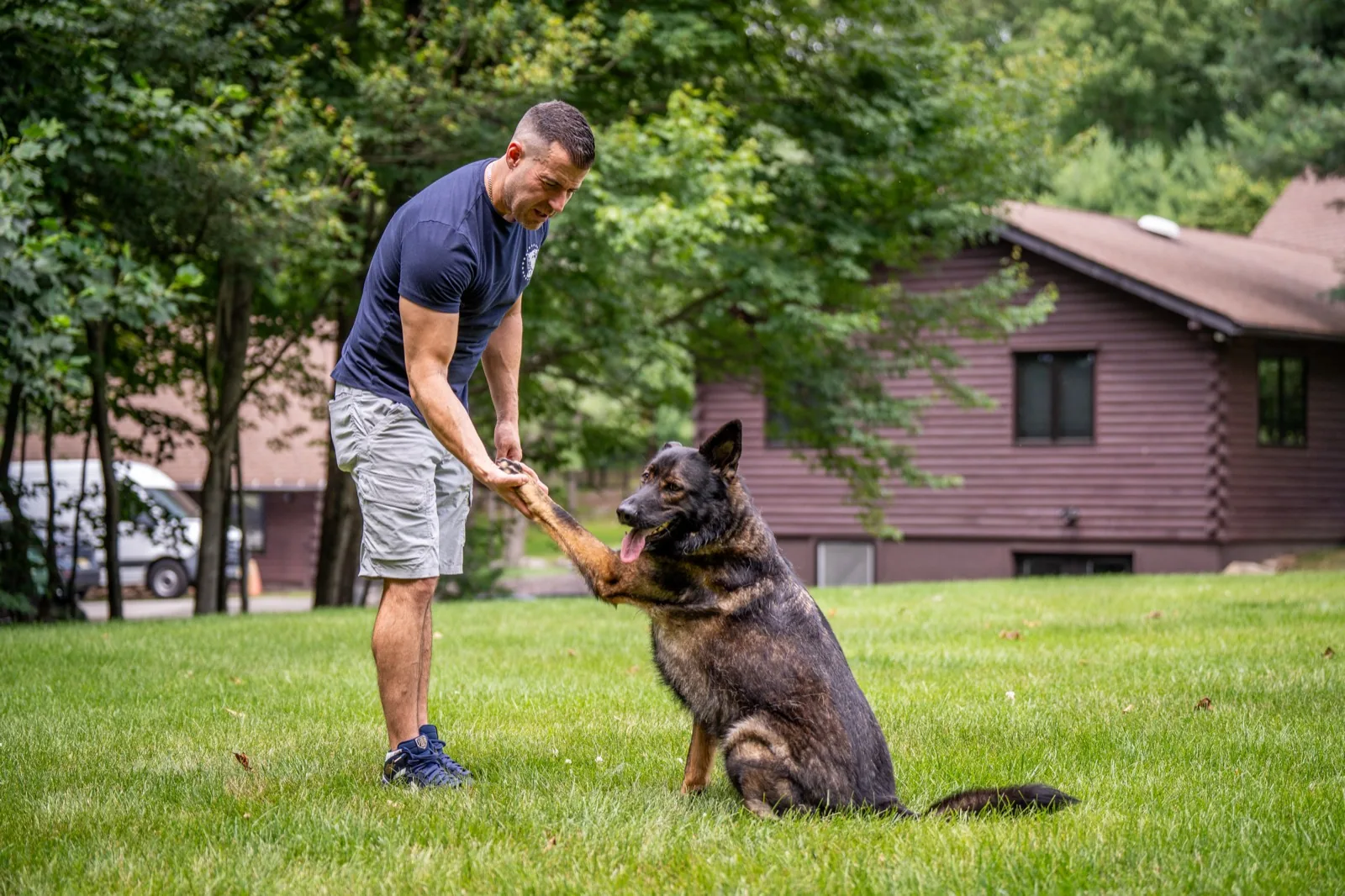 Steve Del Savio taking the offered paw of a seated German Shepherd on the lawn, the lodge behind them.