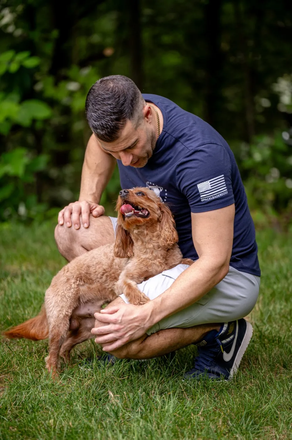 Steve Del Savio kneeling on the lawn with a young cavapoo in his lap, the puppy looking up at his face.