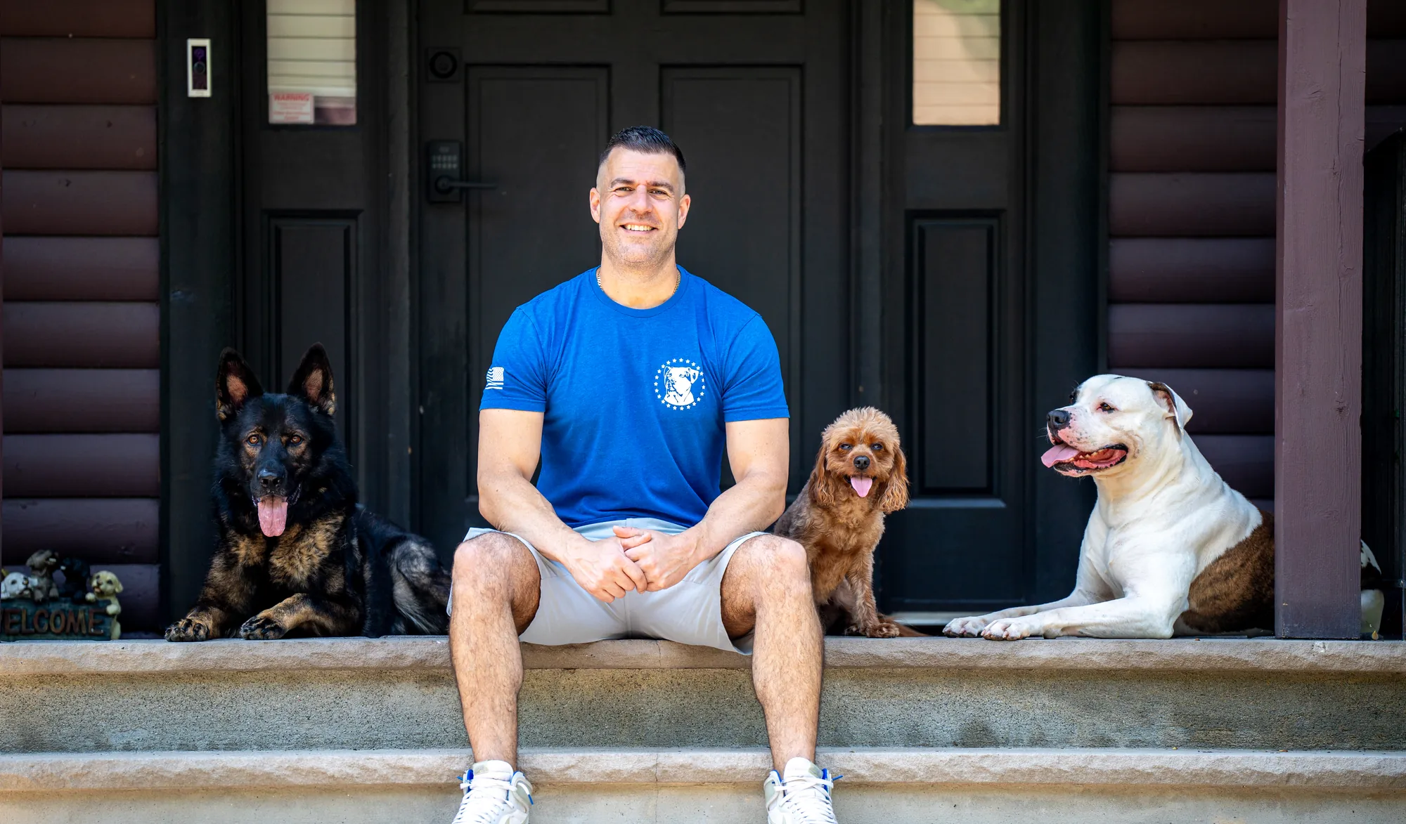 Steve Del Savio seated on the lodge steps with a shepherd, a cavapoo and a bulldog beside him.