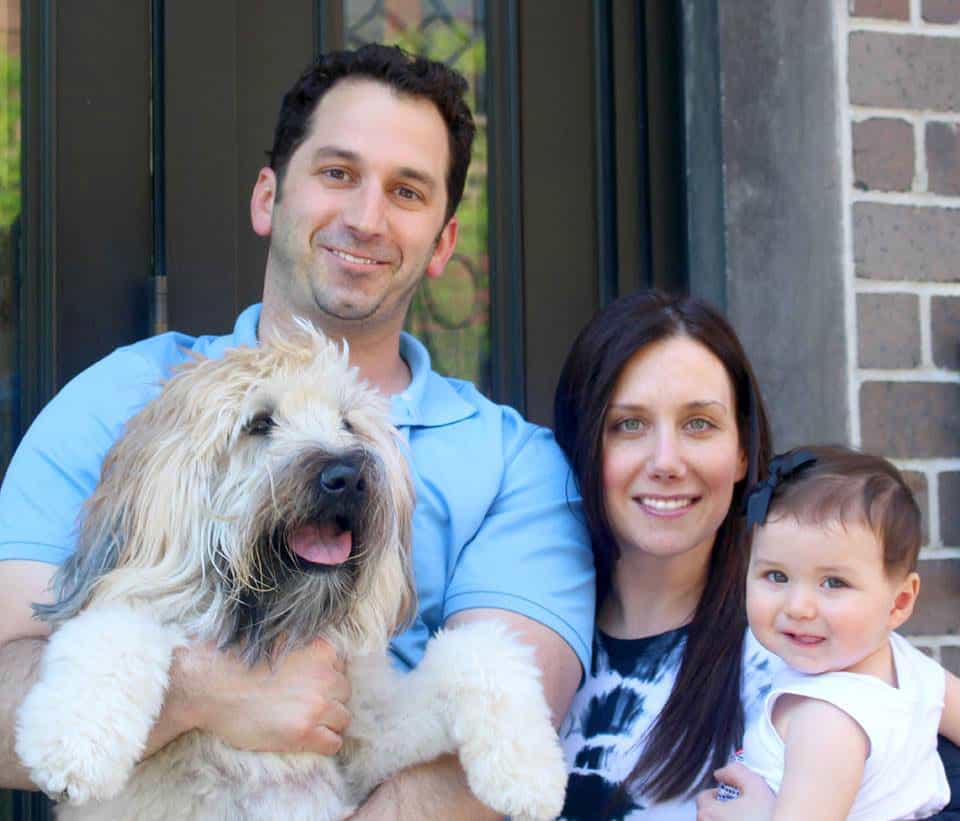 Brooke, Brian and their baby daughter at their front door, Emmitt the wheaten terrier happy in Brian's arms.