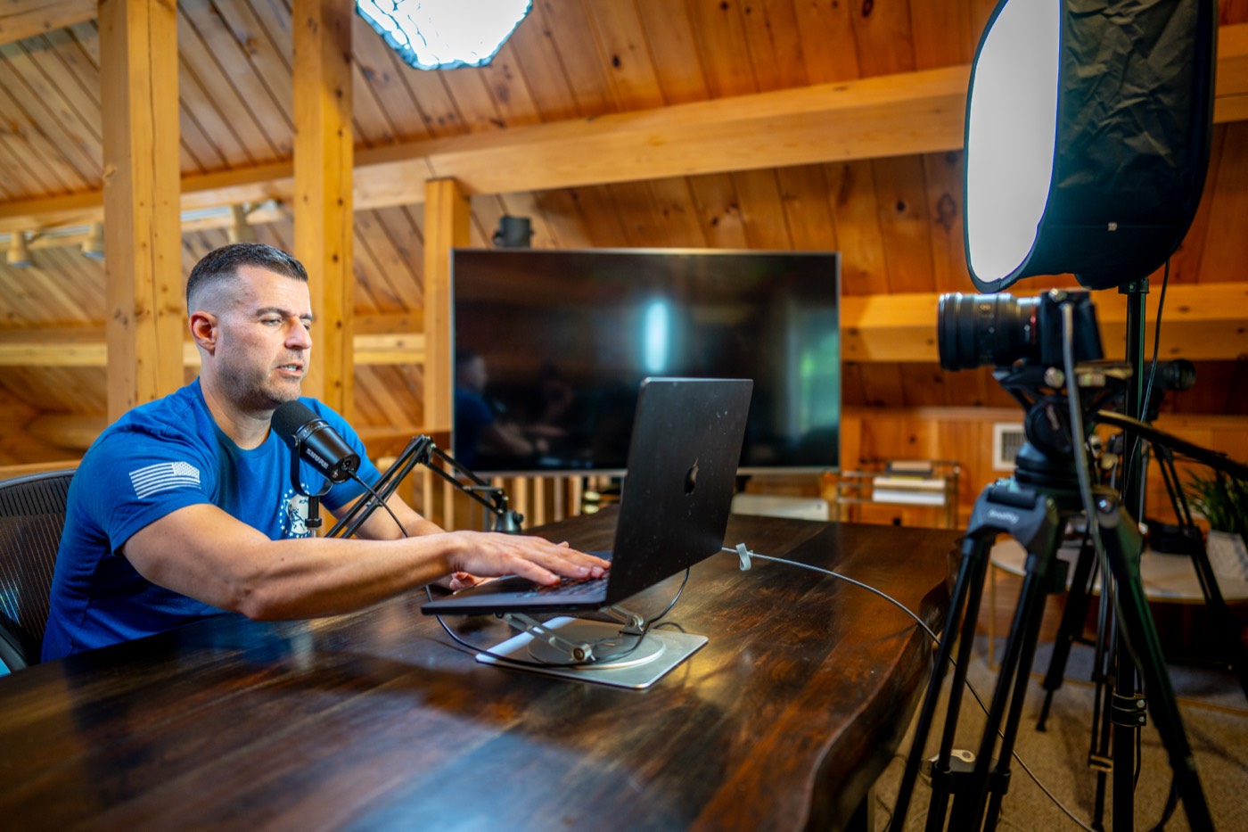Steve Del Savio at a desk in the timber loft of the lodge, speaking to a camera beside a laptop and microphone.